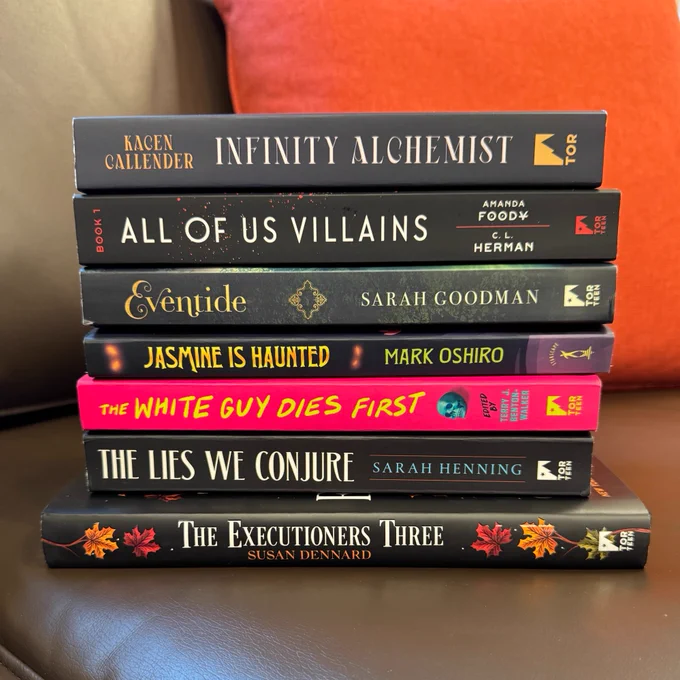 A stack of books standing on a brown leather couch in front of an orange pillow. The books in the stack are, in order from top to bottom: Infinity Alchemist by Kacen Callender, All of Us Villains by Amanda Foody & C.L. Herman, Eventide by Sarah Goodman, Jasmine is Haunted by Mark Oshiro, The White Guy Dies First edited by Terry J. Benton-Walker, The Lies We Conjure by Sarah Henning, and The Executioners Three by Susan Dennard.