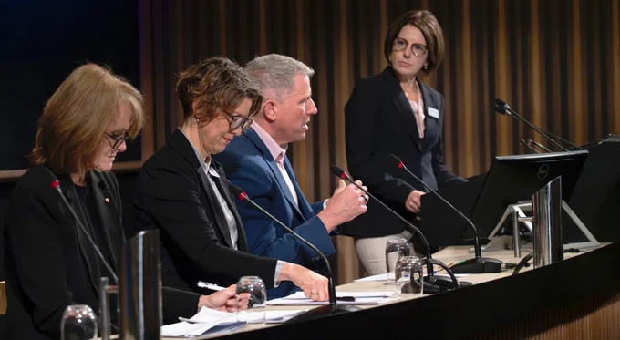 Four professionals at a conference table participating in a panel discussion; two are reviewing notes, one is speaking into a microphone, and another is standing by with a clipboard.