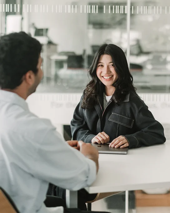 Two students are seated opposite each other at a table in front of a glass wall. The student facing the audience is holding a laptop in their hands, and smiling