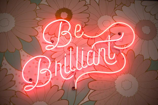 Neon sign that reads "Be Brilliant" in cursive lettering, set against a floral background featuring large daisies.