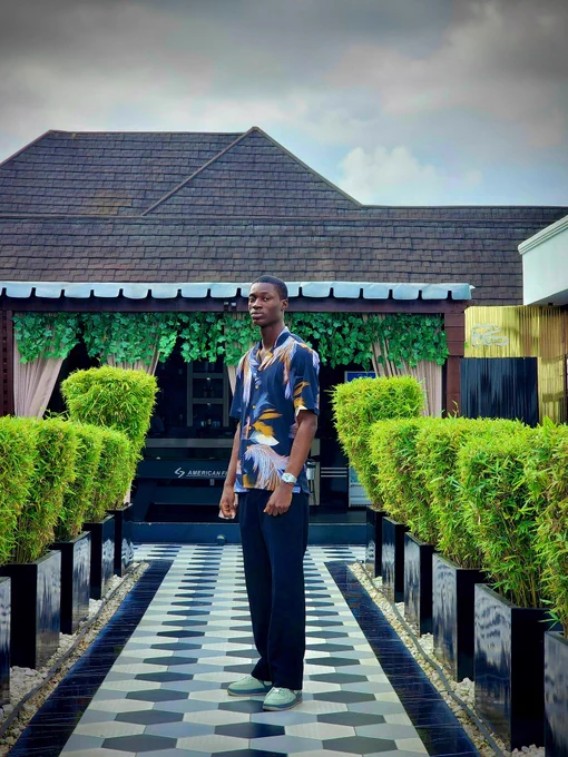 A man stands confidently on a tiled walkway lined with green hedges. He wears a bold short-sleeve button-up shirt with a navy base and gold, white, and blue feather-like patterns from dhukecollectibles, paired with dark trousers and a silver wristwatch. The setting is outdoors with a modern building and greenery in the background, giving off a fresh summer vibe.
