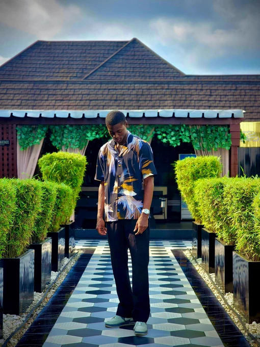 Model in a bold navy short-sleeve shirt with gold, white, and blue feather patterns from Dhuke Collectibles. Standing on a tiled walkway with green hedges and a modern building, capturing a fresh summer vibe.
