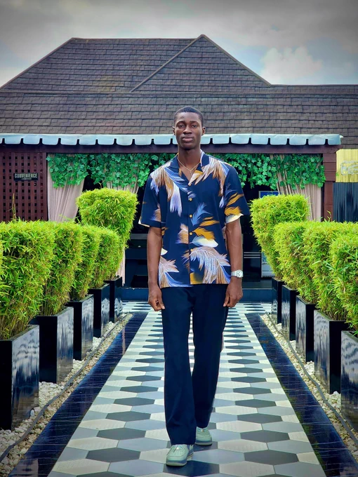 Model in a bold navy short-sleeve shirt with gold, white, and blue feather patterns from Dhuke Collectibles. Standing on a tiled walkway with green hedges and a modern building, capturing a fresh summer vibe.
