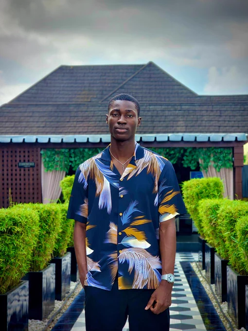 A man stands confidently on a tiled walkway lined with green hedges. He wears a bold short-sleeve button-up shirt with a navy base and gold, white, and blue feather-like patterns from dhukecollectibles, paired with dark trousers and a silver wristwatch. The setting is outdoors with a modern building and greenery in the background, giving off a fresh summer vibe.
