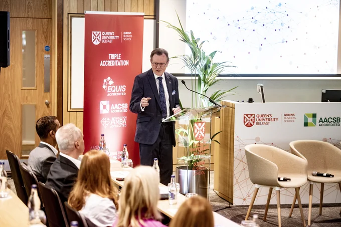 Image shows the President and Vice-Chancellor, Professor Sir Ian Greer, speaking at an event to mark the triple accreditation. Behind him is a promotional stand with the Queen's Business School logo and the logos of the three accreditations.