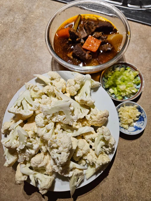 Ingredients from top of a bowl of slowcooked beef, daikon, carrot, lemongrass, a small dish of spring onion slices, a small dish of minced garlic, a plate of cauliflower pieces.