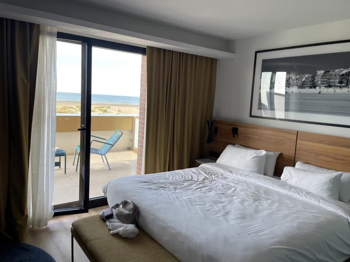 big hotel bed in the foreground, a glassdoor and a balcony with sea view in the background