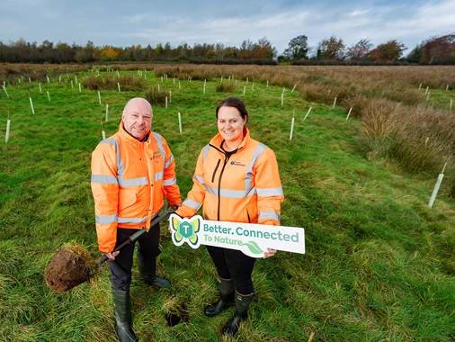 Male and female in a field, both are wearing orange hi-vis jackets and the female is holding a sign that says " Better Connect To nature" with the Translink logo
