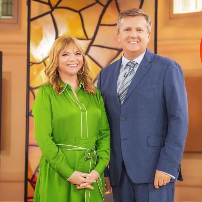Rev. Kate Bottley and Aled Jones stood next to each other in front of some stained glass windows, smiling. The Big School Assembly Singalong logo is in a red circle at the top right of the image