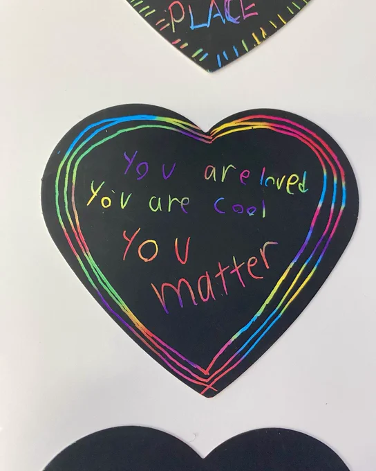 Black heart-shaped paper with a rainbow-colored border and the handwritten message: 'You are loved, You are cool, You matter' in multicolored letters.