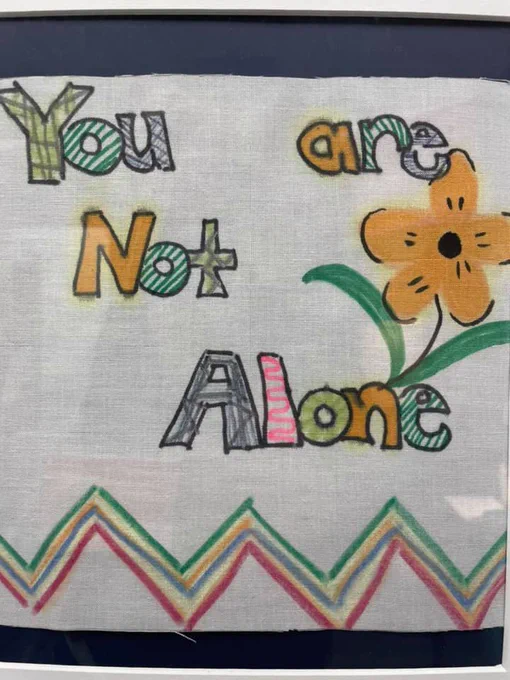 Hand-drawn colorful text on fabric reads 'You are Not Alone' with a large orange flower and green leaves beside the words.