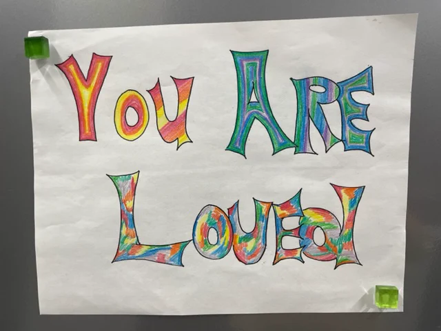 Hand-drawn sign with colorful, stylized letters spelling out 'You Are Loved' pinned to a gray surface with green magnets.