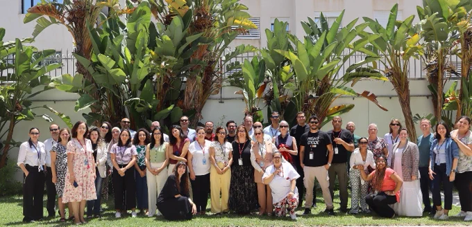 A large group of people posing outdoors on a sunny day. They are standing in front of some palm trees and the exterior wall of a white building.