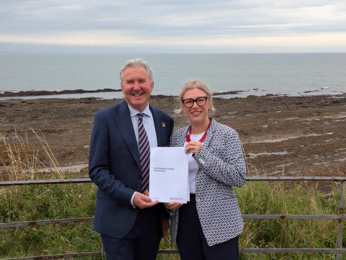 Professor Bill Austin  and Climate Action Secretary Gillian Martin hold a copy of the Scottish Government's Blue Carbon Action Plan with salt marsh and sea in the background.