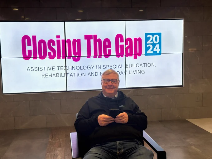 A smiling man sits in front of a large digital screen displaying the words ‘Closing The Gap 2024 – Assistive Technology in Special Education, Rehabilitation and Everyday Living.’ He is holding a phone and wearing glasses and a black sweatshirt.