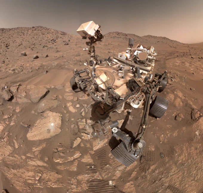 A self-portrait photo of the Perseverance rover on the surface of Mars, taken with a camera on its robotic arm. The six-wheel rover sits in a rocky desert landscape. Its robotic "head" is pointed down to one rock where there is a small hole drilled, along with a small pile of light-colored tailings from the drill. 
