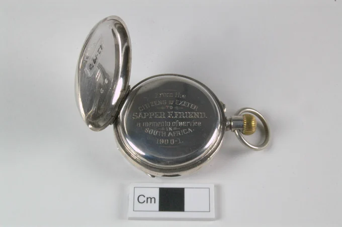 Back of a silver pocket watch with the lid open, revealing an engraved inscription: "From the citizens of Exeter to Sapper K. Friend, a memento of service in South Africa 1900-1." A centimeter scale is placed below for size reference.