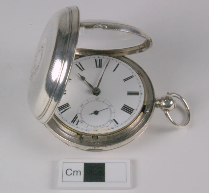 Open silver pocket watch displaying a white dial with black Roman numerals and a small seconds subdial, placed beside a centimeter scale.