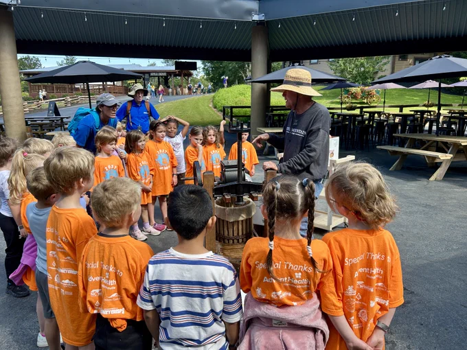 group of students in orange class of 2037 shirts gather around a man at a cider press in a half circle. he is wearing a jeter mountain farm shirt and wearing a hat.
