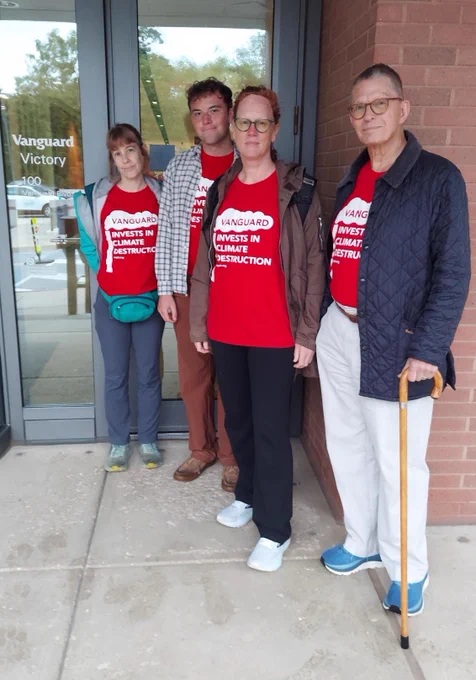 [Alt text: People people in matching red shirts pose for the camera in front of a building that says “Vanguard Victory.” Photo by Lee Smithey ] 
