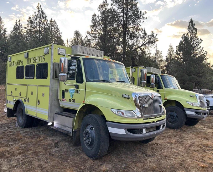 Two light yellow firetrucks are parked near a forest.