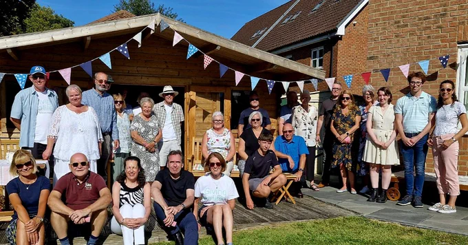 A large group of just under 30 people gathered in a garden. The weather is sunny, and there is bunting across a large shed which they are gathered in front of.