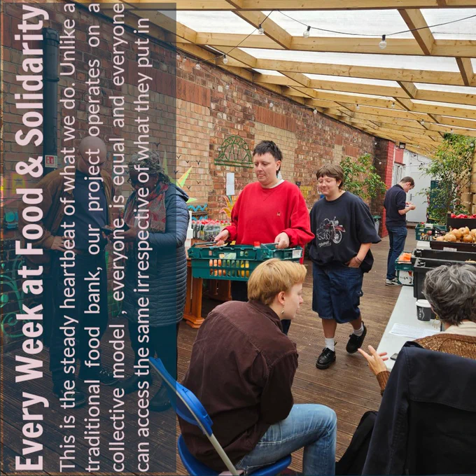 Inside a community food space with wooden beams and brick walls, people are gathered around tables of food. A person in a red jumper carries a green crate of fruit and vegetables, while others chat and sit nearby. The text along the left side reads: “Every Week at Food & Solidarity – This is the steady heartbeat of what we do. Unlike a traditional food bank, our project operates on a collective model, everyone is equal and everyone can access the same irrespective of what they put in.”