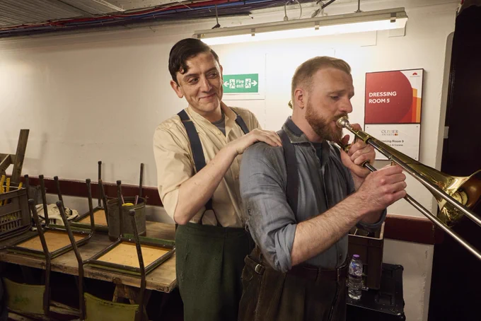 Two musicians backstage, one tuning a trombone while the other watches, surrounded by musical equipment and stage props, with signs including "Dressing Rooms" in the background.