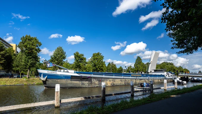 A large cargo ship named VAARWEL, painted in blue and white, is sailing through a canal. The ship has a flag at its front and is passing under an open drawbridge in the background. The canal is lined with trees on both sides, and the sky above is bright blue with scattered white clouds. A few people are visible on the bridge and near the water’s edge. In the foreground, at the bottom right, a small boat with a single person inside floats near the ship.