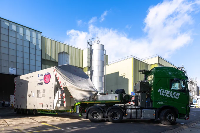 A large green truck from Kübler Spedition is transporting a massive container wrapped in a protective tarp. The container displays several logos and text, including the logo of the European Space Agency (ESA), indicating it is part of a specialised transport operation. In the background, there is an industrial building with large metal silos under a blue sky.