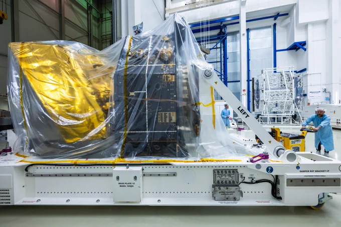 In a cleanroom environment, two individuals wearing blue lab coats and hairnets are working around a large spacecraft. The spacecraft is covered in protective plastic sheeting and mounted on a white transport platform labelled with technical instructions.