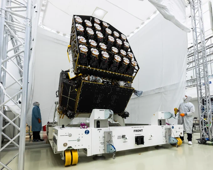 In a cleanroom environment, a group of engineers wearing protective coats, gloves, and face masks are working on a large spacecraft component. The component is mounted on a white wheeled platform. The top of the spacecraft carries 26 cylindrical cameras. The workspace is surrounded by structural supports and white fabric walls, creating a clean and controlled environment.