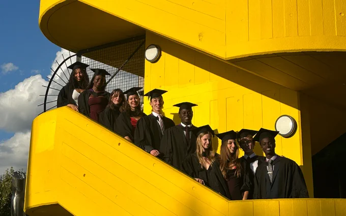 Group of graduates in caps and gowns, posing on the yellow stairs of the Southbank Centre.