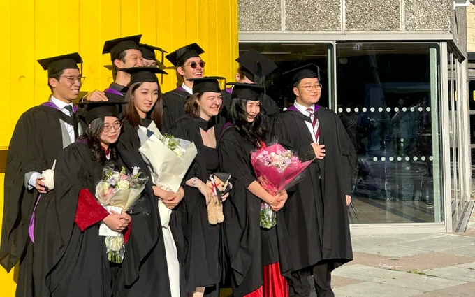 Group of graduates in caps and gowns holding diplomas and flowers, smiling during a sunny graduation ceremony.