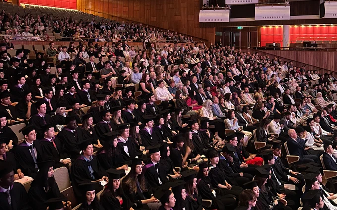 A large crowd of graduates in black robes and caps seated at a graduation ceremony inside a spacious auditorium.