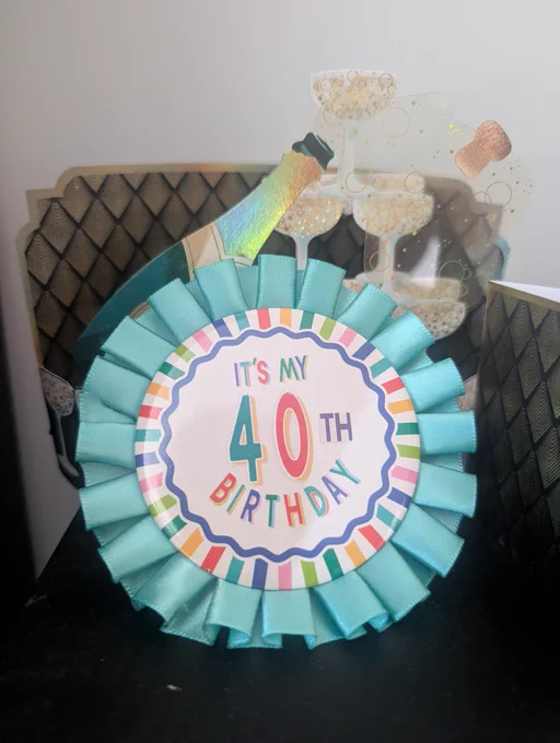 The photo shows a colorful birthday badge with a teal ribbon edge and a round center that reads: “IT’S MY 40TH BIRTHDAY”. Behind it there’s a decorative pop-up design of a champagne bottle popping and bubbly drinks.