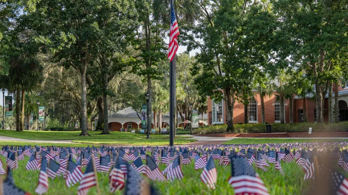 A view of a park with 2,977 American flags placed on the ground, trees in the background, and a Stetson University building with brick facade visible.