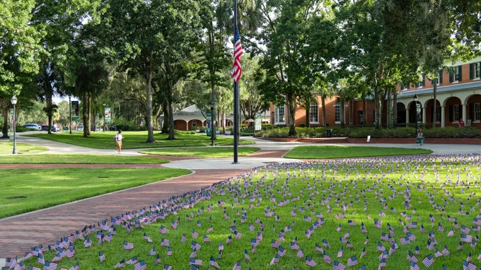 A view of Stetson University's campus, showing walkways lined with 2,977 American flags, a green lawn, and classical buildings in the background under a clear sky. An American flag flutters prominently on a pole.