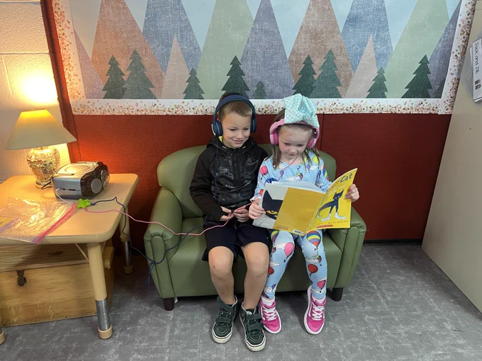 two kindergarten students sit on a couch and listen to a read aloud of pete the cats with their headphones
