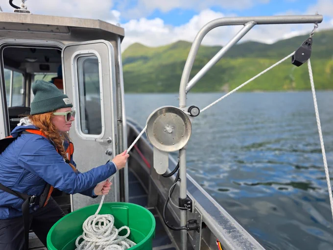 woman on ship pulls on white rope through pulley with ocean and mountains visible in the background 