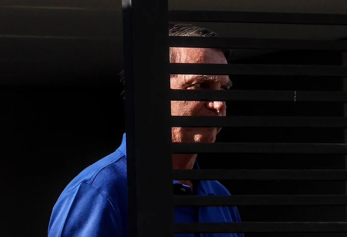 Black and white photo by Gabriela Biló showing Jair Bolsonaro in profile, looking down with a somber expression. He wears a suit, tie, and the presidential sash. Horizontal bars in the foreground give the impression that he is behind bars, but they are actually part of the gate at the house where he is under house arrest.
