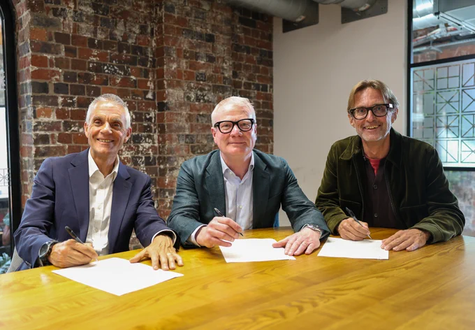 Photograph of three men sat down at a table each signing a document with a pen.  