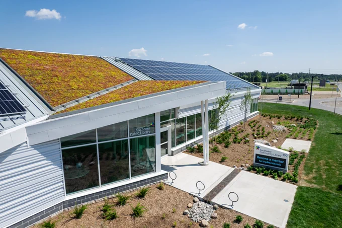 Modern community library building with landscaping, green roof sections, and solar panels on a sunny day. 