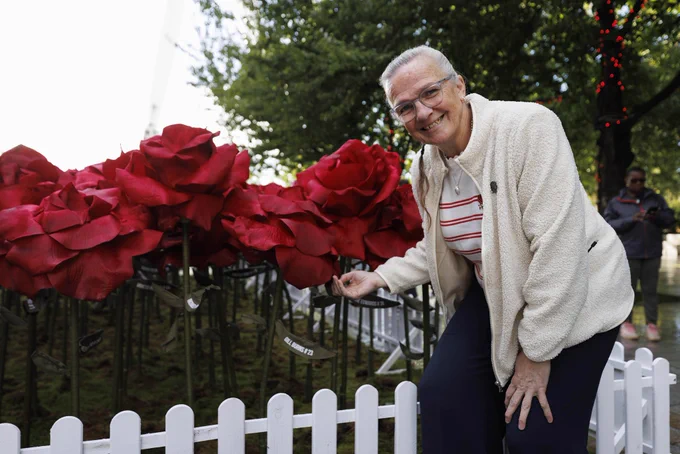 Gill Burns posing in front of a handmade red rose with her name and legacy number on it.