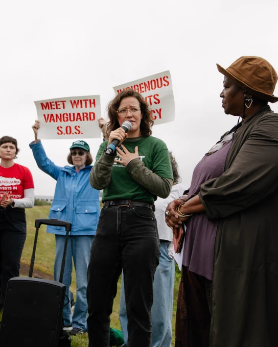 A person speaks into a microphone while looking at another person, and others stand around them holding signs. Photo by Rachael Warriner
