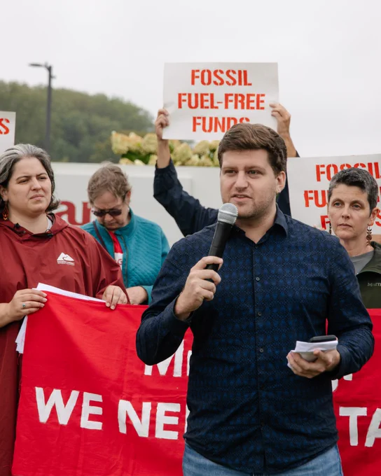 A person speaks into a microphone while others stand behind them, holding banners and signs, including one that says “fossil fuel-free funds.” Photo by Rachael Warriner