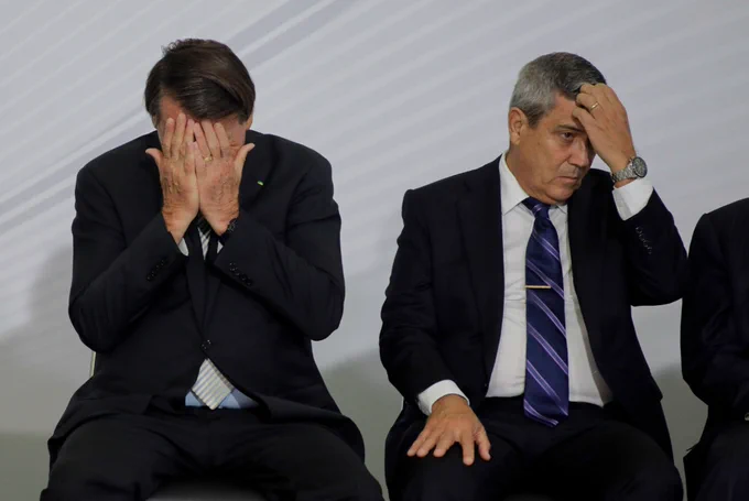 Jair Bolsonaro (left) covers his face with both hands in a gesture of frustration or despair, while General Walter Braga Netto (right), a four-star general, sits beside him with a tense expression, touching his forehead. Both are dressed in dark suits and seated against a plain background, conveying a somber or stressful moment.