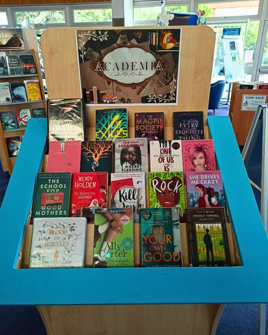  Photograph of a large wooden book display with tiered shelves. A poster at the top of the display reads "Academia". Books on the display include All The Devils by Catelyn Wilson, Every Exquisite Thing by Laura Steven, Solitaire by Alice Oseman, One of Us is Lying by Karen McManus, The School For Good Mothers by Jessamine Chan, Blood of Hercules by Jasmine Mas, and Summer Half by Angela Thirkell.