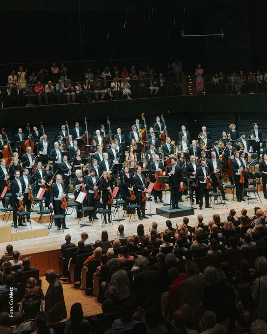 Dirigent Lahav Shani mit den Münchner Philharmonikern auf der Bühne , stehend vor dem applaudierenden Publikum.