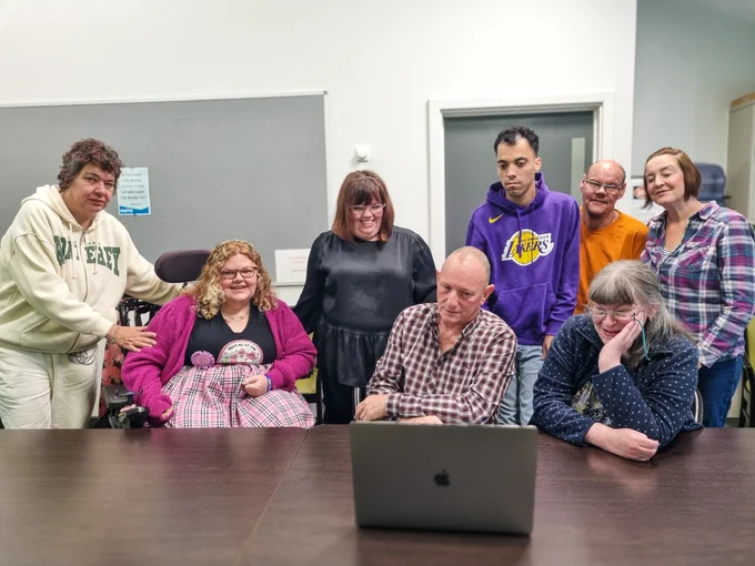 Dave (Lippy People) is sat with a group of people, with mixed abilities and of all backgrounds, who are watching a laptop, as they review a video.
Some people are smiling in fond memory, some people look sad and some people look serious. 